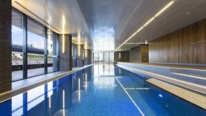 Piscina interior moderna com paredes de vidro transparente, chão de azulejos azuis e parede de madeira de um lado.