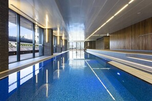 Una piscina interior moderna con paredes de vidrio transparente, suelo de baldosas azules y una pared de madera en un lateral.