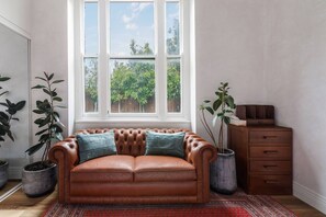 A brown leather sofa with two cushions, a window with a view of trees, and a wooden dresser with drawers.