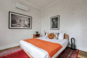 A bedroom with a bed, a red rug, a small bedside table with a plant, and two framed pictures on the wall.