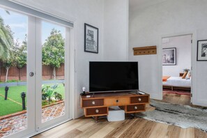 Un salon moderne avec un téléviseur à écran plat posé sur un meuble en bois, une porte-fenêtre coulissante donnant sur un jardin et un lit surmonté d’un tapis rouge.