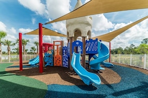 A playground with multiple slides and climbing structures, surrounded by a fence and palm trees. - Solara Resort Villa-9BR | Pool & Spa | Near Disney (Kissimmee)