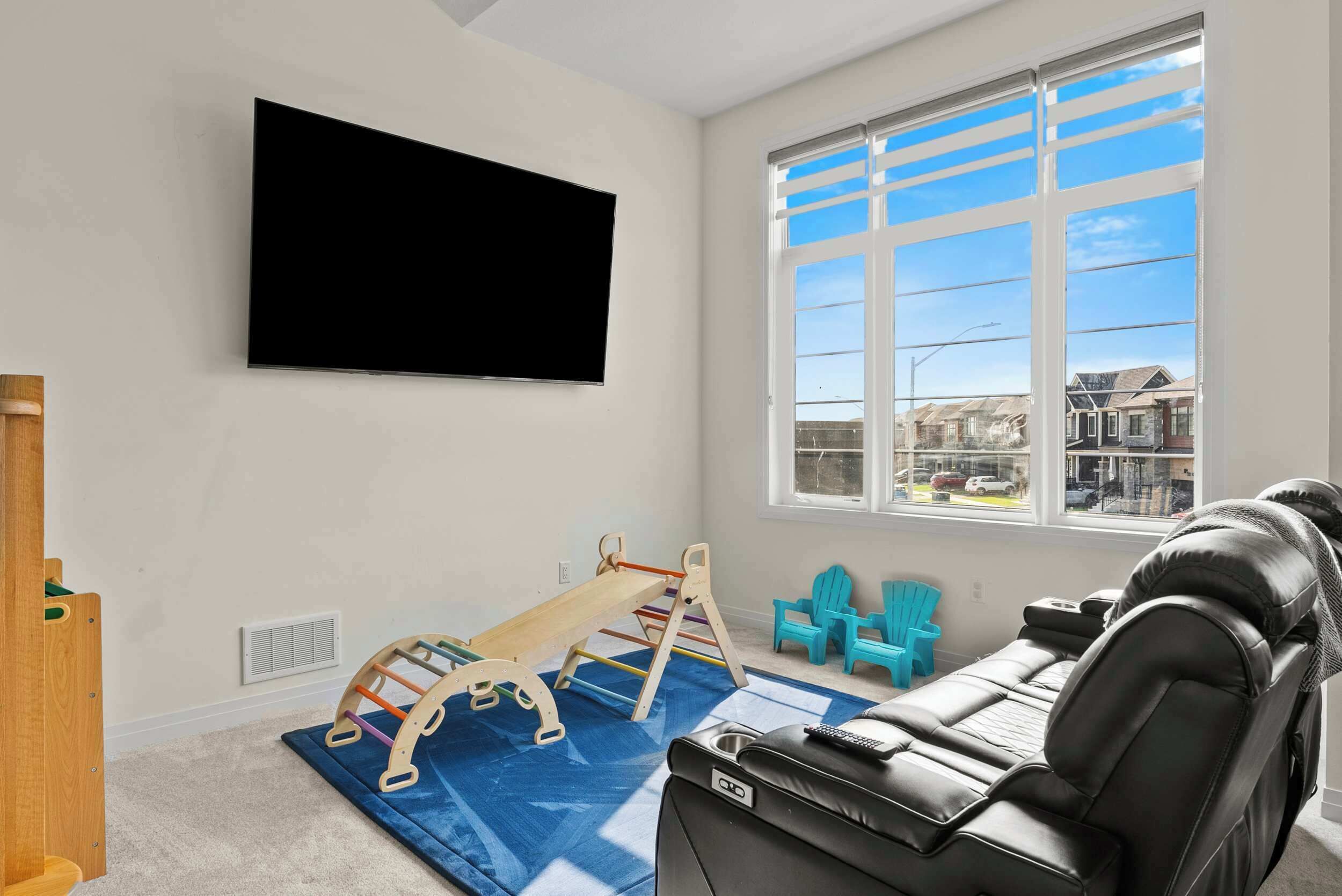 A room with a mounted TV, a window with a view of houses, a playpen, and a sofa.