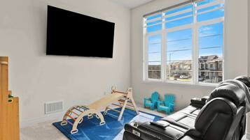 A room with a mounted TV, a window with a view of houses, a playpen, and a sofa.
