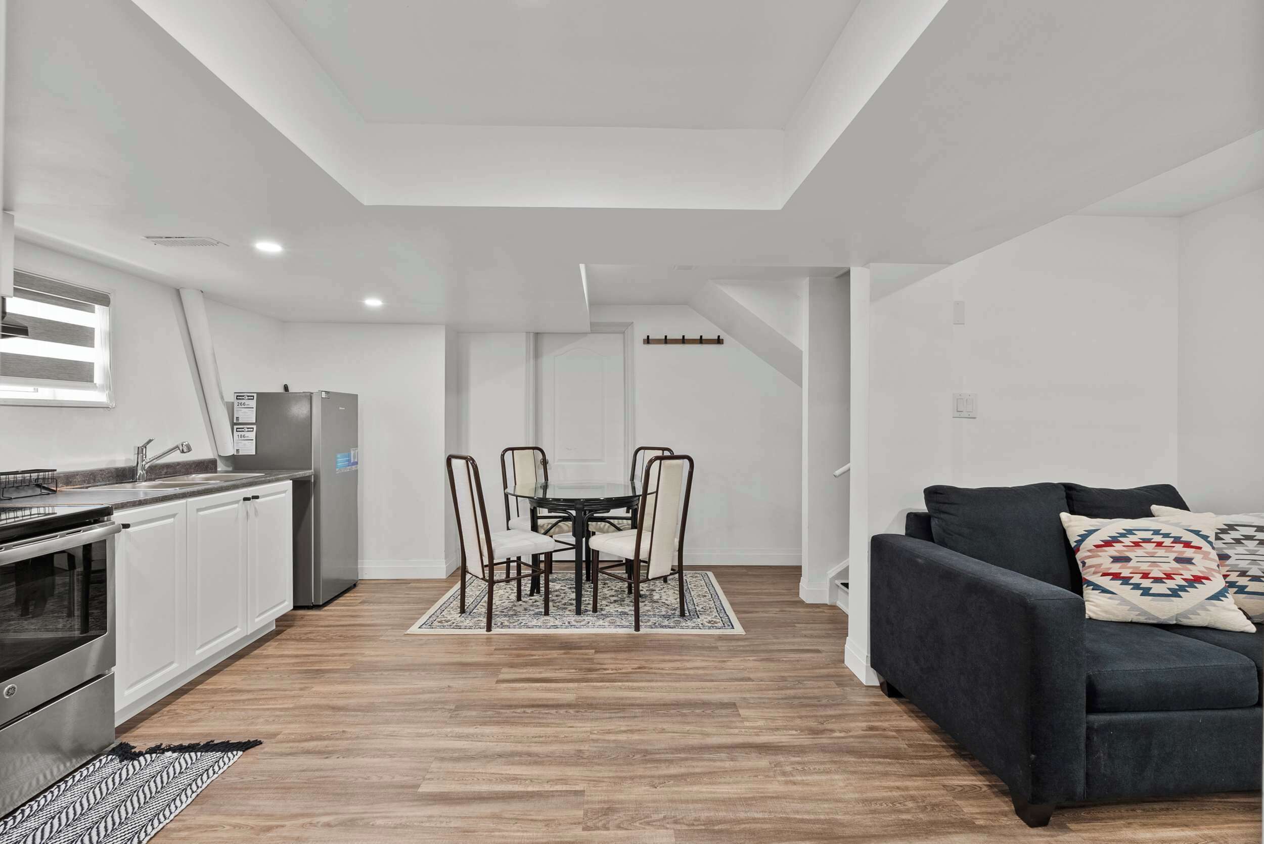 A modern kitchen with a built-in oven, a dining area with a glass table and metal chairs, and a dark-colored sofa with patterned cushions.