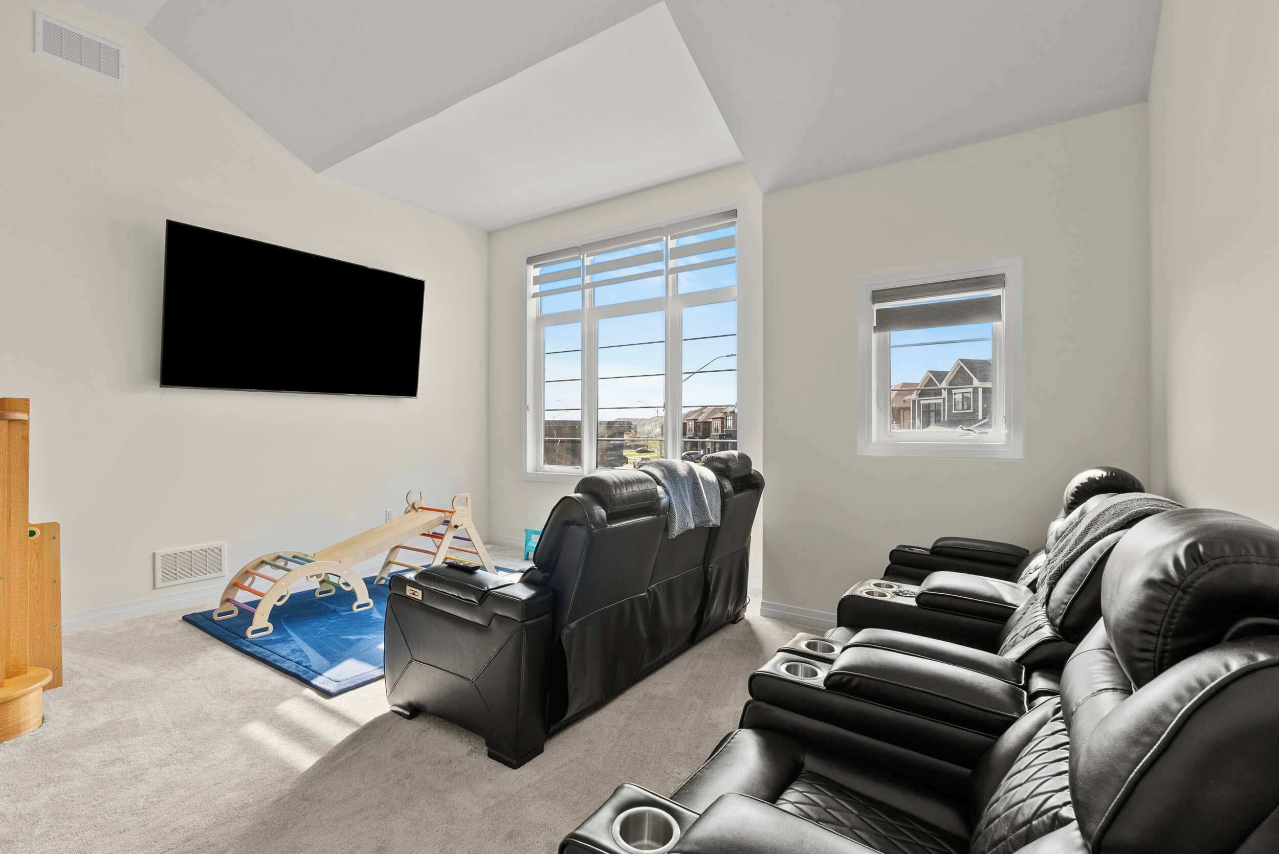 A living room with a flat-screen TV, a massage chair, a wooden play structure, and a view of houses through the windows.