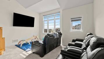 A living room with a flat-screen TV, a massage chair, a wooden play structure, and a view of houses through the windows.