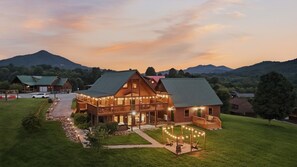 A large wooden lodge with multiple balconies and string lights, surrounded by a well-maintained lawn and mountains in the background. - Overwatch: HUGE Game Room, Hot Tub, FirePit, &More (Pigeon Forge)