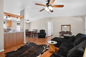 Amplia sala de estar con un sofá de cuero negro, una mesa de comedor de madera con sillas, una zona de cocina con armarios de madera y un ventilador de techo.