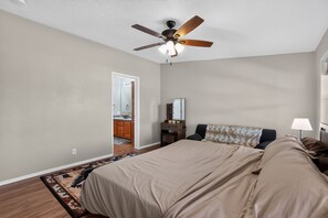 A bedroom with a bed, a nightstand, a lamp, a ceiling fan, and a bathroom visible through an open door. - Cozy AZ Desert Escape – Hiking, OHV & Mtn Views (Black Canyon City)