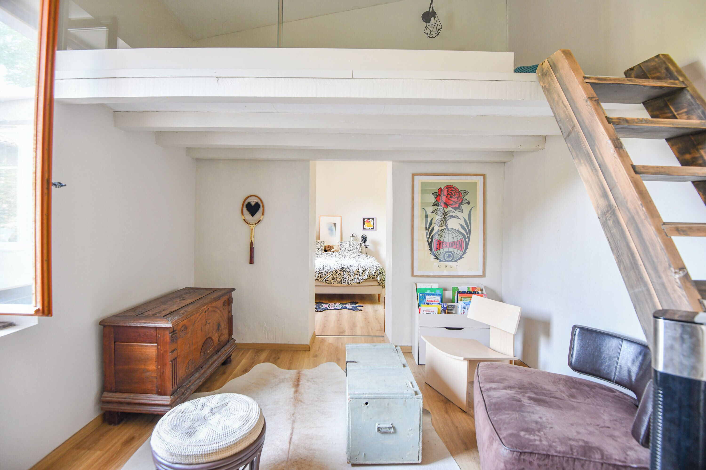 A cozy room with a wooden chest, a small round table, a cushioned bench, a metal box, and a ladder leading to a loft area.
