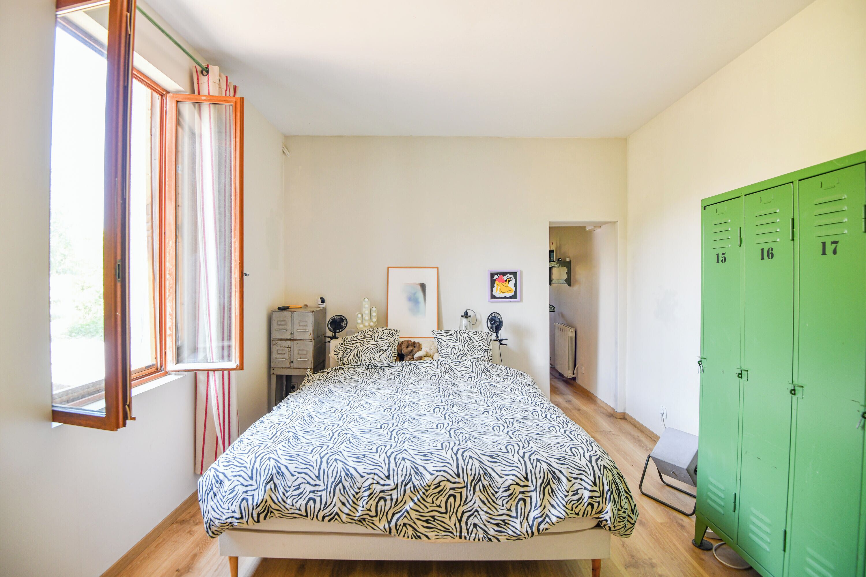 A bedroom with a bed, a window, a green wardrobe, and a door leading to another room.