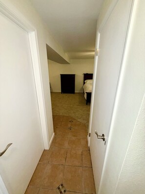 A narrow hallway with a carpeted floor leading to a bedroom with a bed and a dresser.