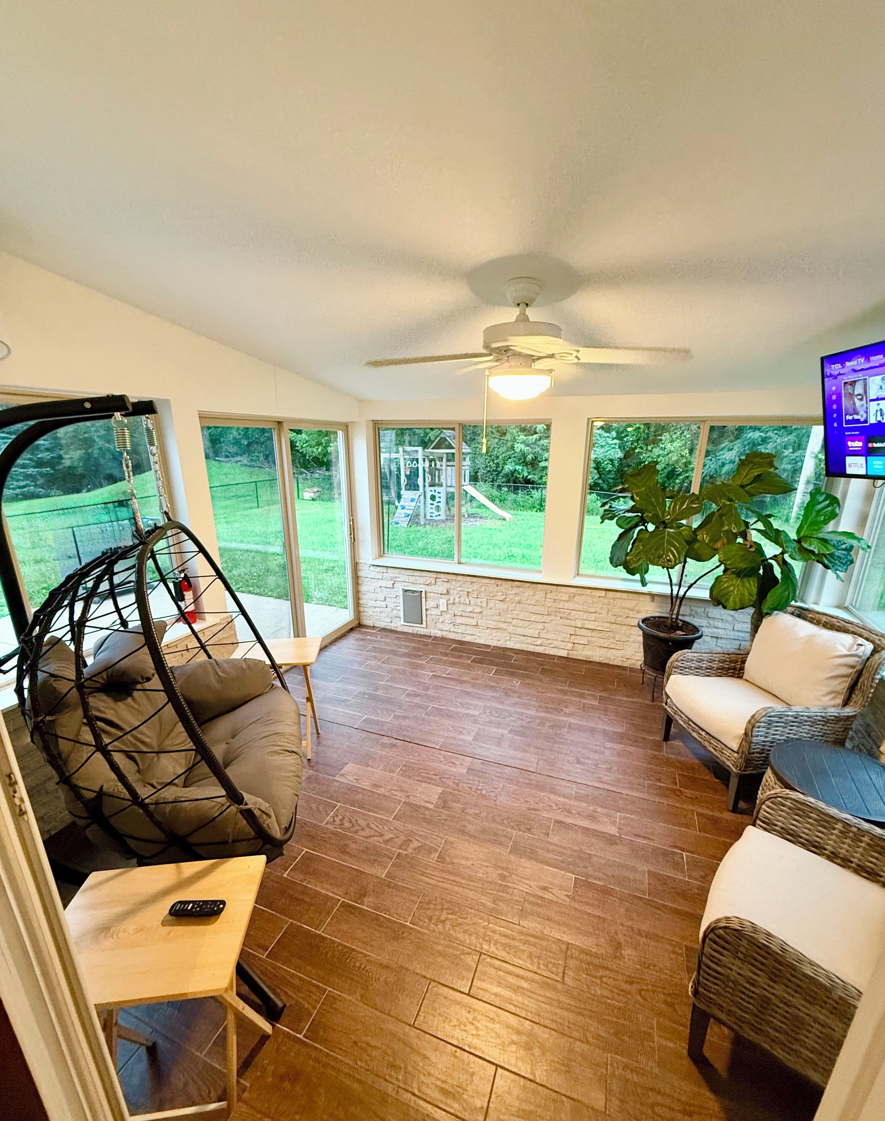 A spacious room with a ceiling fan, large windows, a swing chair, and a television.