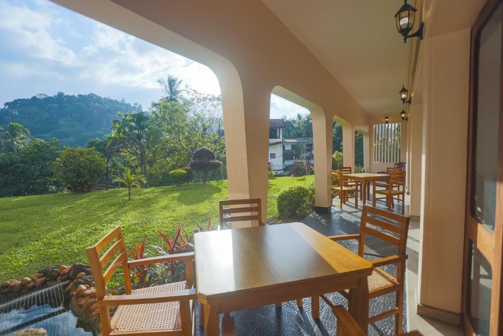 A dining area with a table and chairs, a view of a lush garden, and a building in the background.