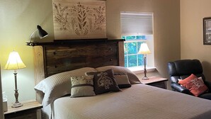 A bedroom with a bed, a chair, a nightstand with a lamp, a ceiling fan, and a window with blinds.