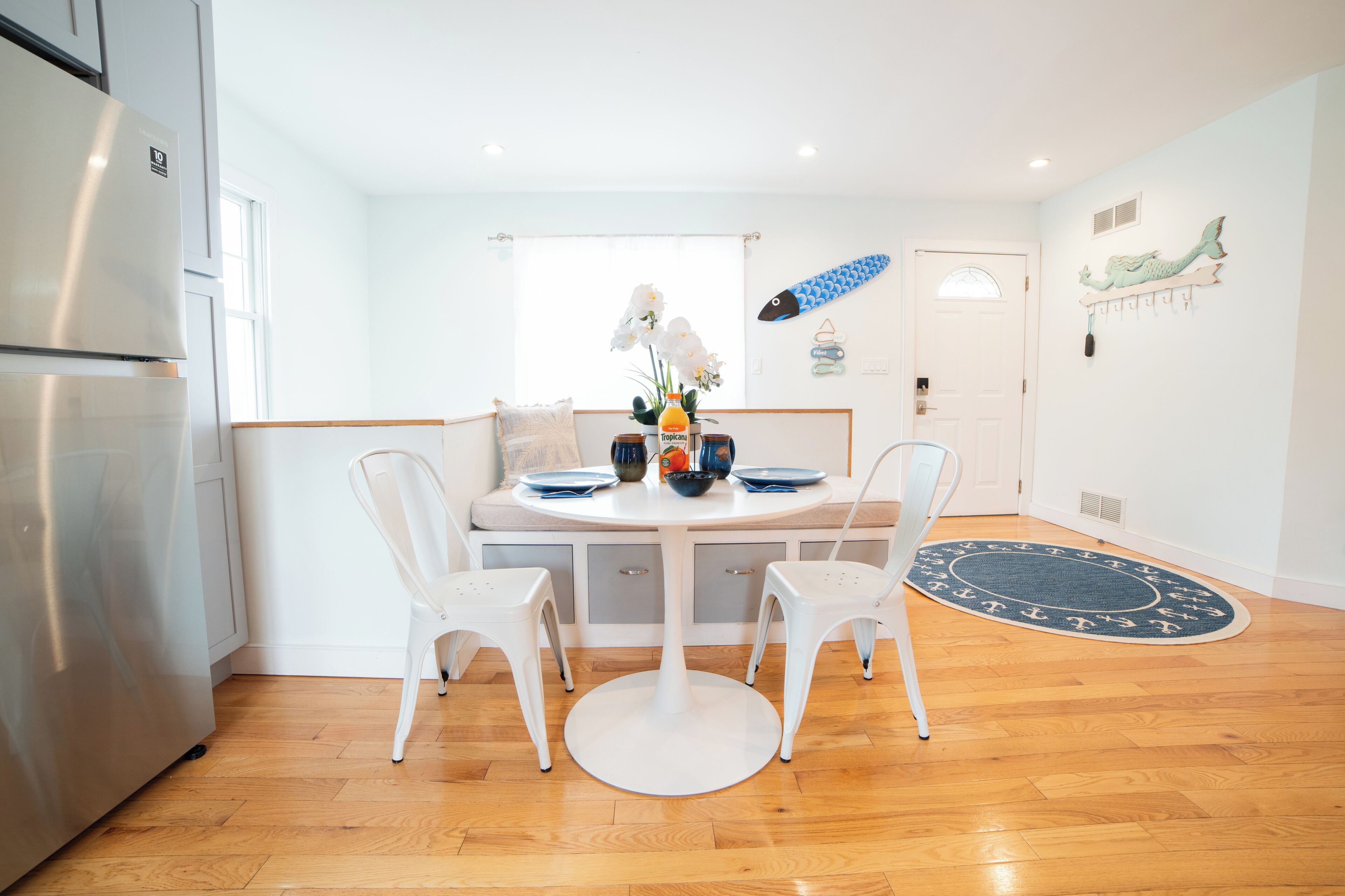 A modern kitchen with white cabinetry, a stainless steel refrigerator, a round dining table with two white chairs, and a blue decorative rug on the floor.