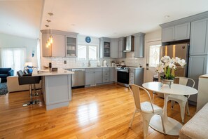 A modern kitchen with grey cabinets, a central island, and a dining area with a white table and chairs. - Hampton Bays Resort-Style Retreat with Pool, Hot Tub, Basketball & Pickleball! (Hampton Bays)