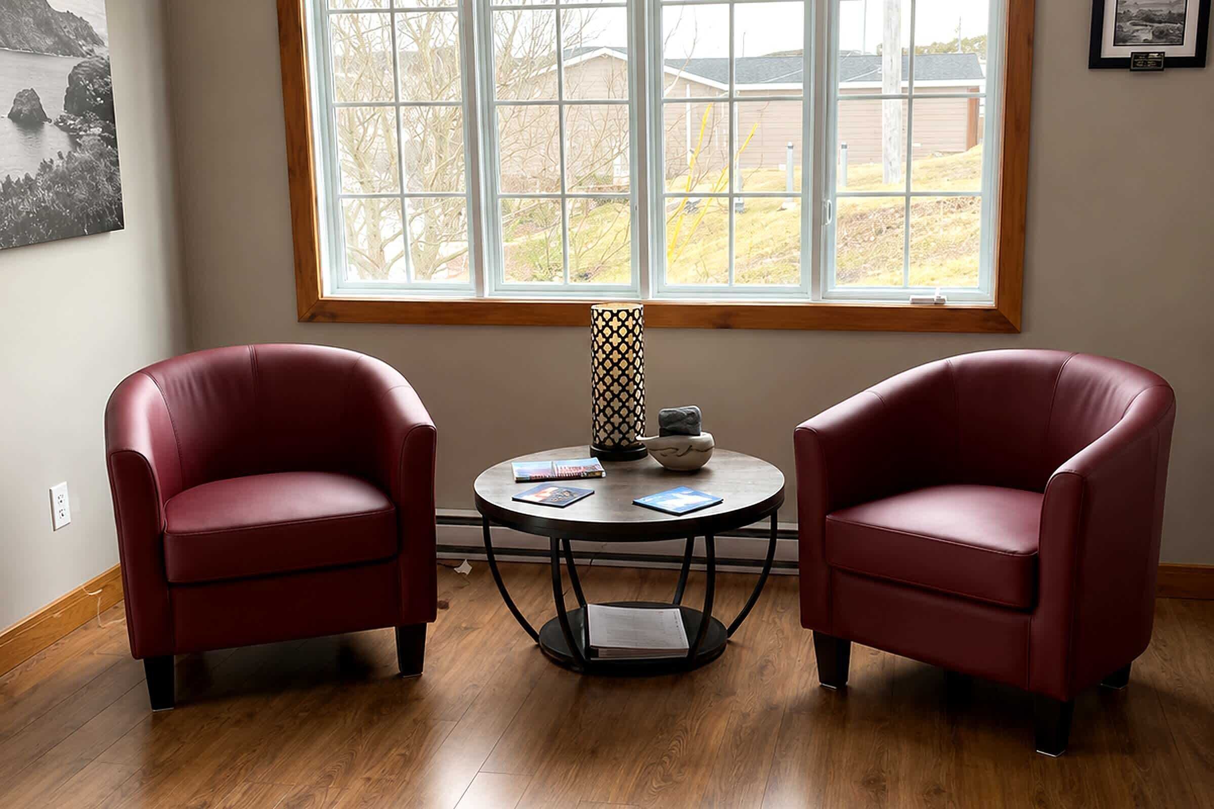 A room with two red leather chairs, a small round table with a book and a decorative item, and a large window with a view of houses and trees.