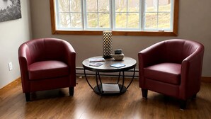 A room with two red leather chairs, a small round table with a book and a decorative item, and a large window with a view of houses and trees.
