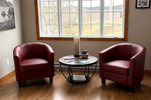 Una habitación con dos sillones de cuero rojo, una mesita redonda con un libro y un adorno, y un ventanal con vistas a casas y árboles.