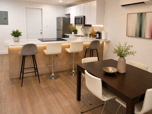 A modern kitchen with white cabinets, a stainless steel refrigerator, a microwave, and a coffee maker. There are three bar stools and a dining table with chairs. - Vrbo Property (North Miami)