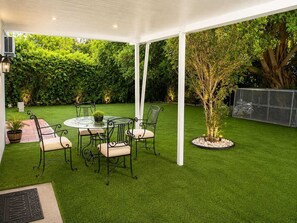 A covered patio with a round table and chairs, surrounded by a well-maintained lawn and a hedge. - Vrbo Property (North Miami)
