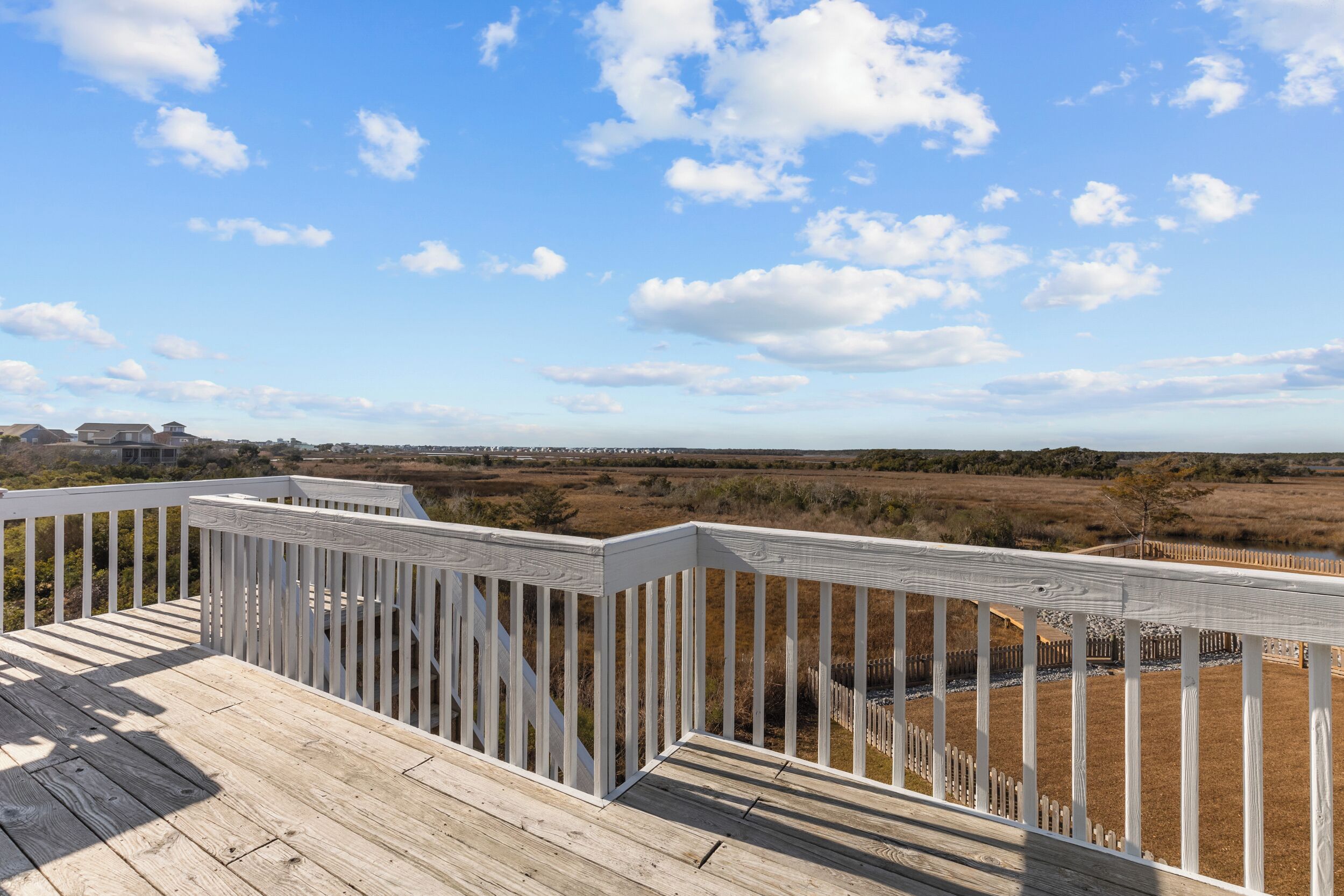 A wooden deck with a white railing overlooking a landscape with a clear sky and distant buildings.