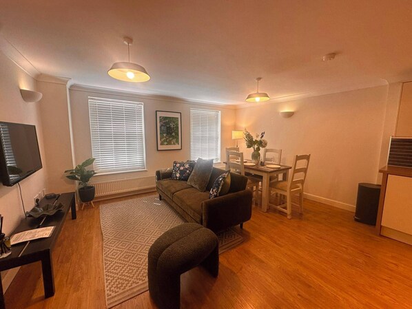 A living room with a sofa, a dining table with chairs, a TV mounted on the wall, and a potted plant. - 2-Bed Flat in Shoreditch, Heart of Brick Lane (London)