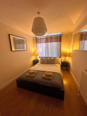 A hotel room with a large bed, two bedside tables with lamps, a ceiling light, a framed picture on the wall, and a radiator. - 2-Bed Flat in Shoreditch, Heart of Brick Lane (London)