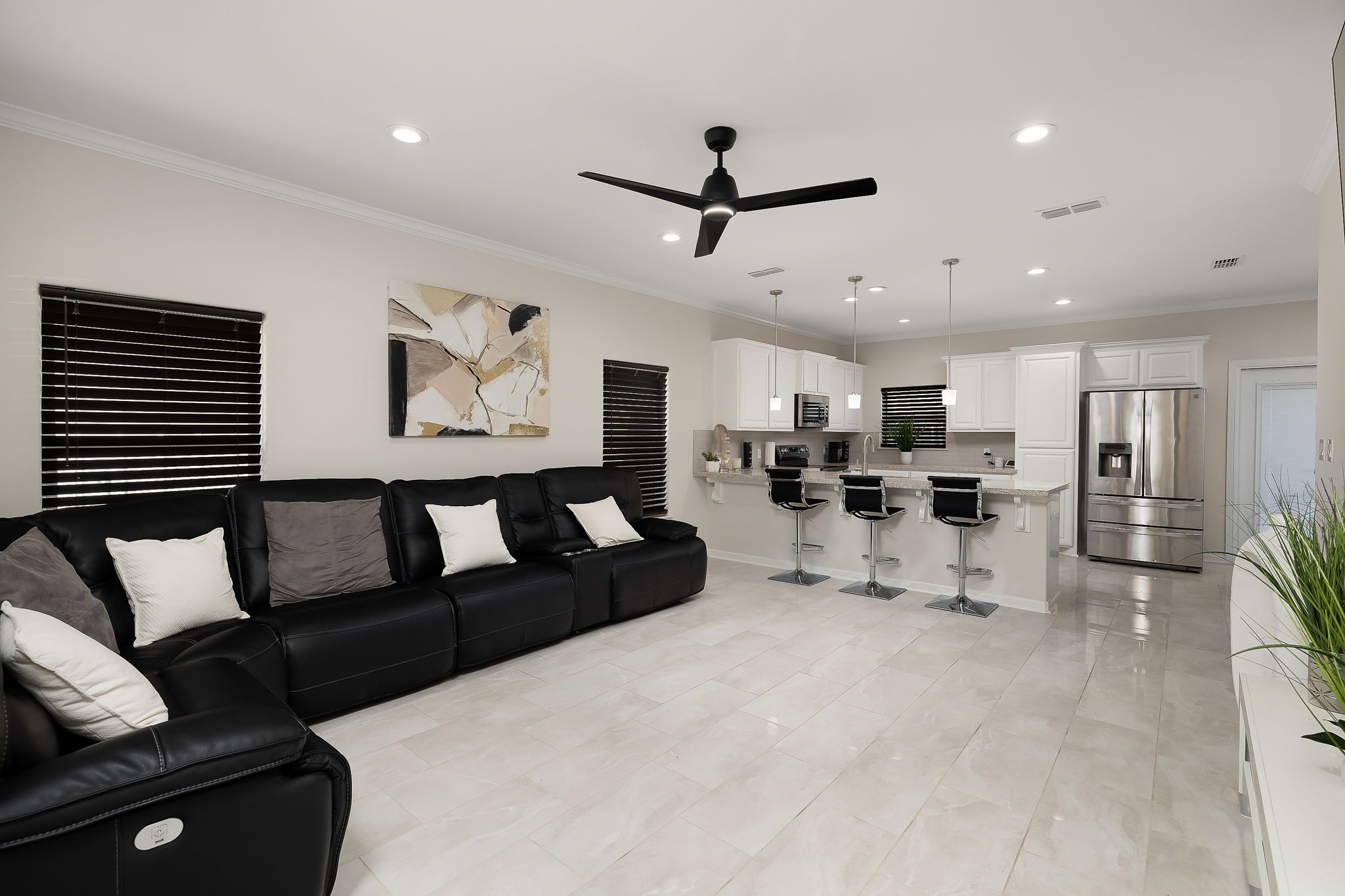 Uma sala de estar moderna com um sofá de couro preto, ventilador de teto e área de cozinha com armários brancos e eletrodomésticos de aço inoxidável.