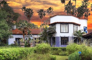 A two-story building with a white exterior and red-tiled roof, surrounded by lush greenery and palm trees.