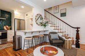 A modern kitchen with a stainless steel refrigerator, a bar area with stools, a staircase with a wooden railing, and a dining area with a round table.