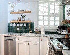 A kitchen with a dishwasher, oven, and a coffee maker. There are wooden shelves above the counter with various items.