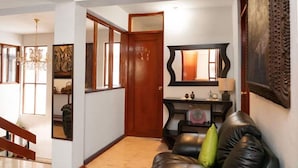 Un pasillo con una puerta de madera, un sofá de cuero negro con un cojín verde, un espejo y un reloj fijado a la pared.