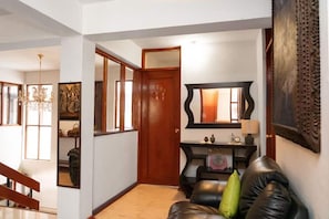 A hallway with a wooden door, a black leather sofa with a green pillow, a mirror, and a wall-mounted clock.