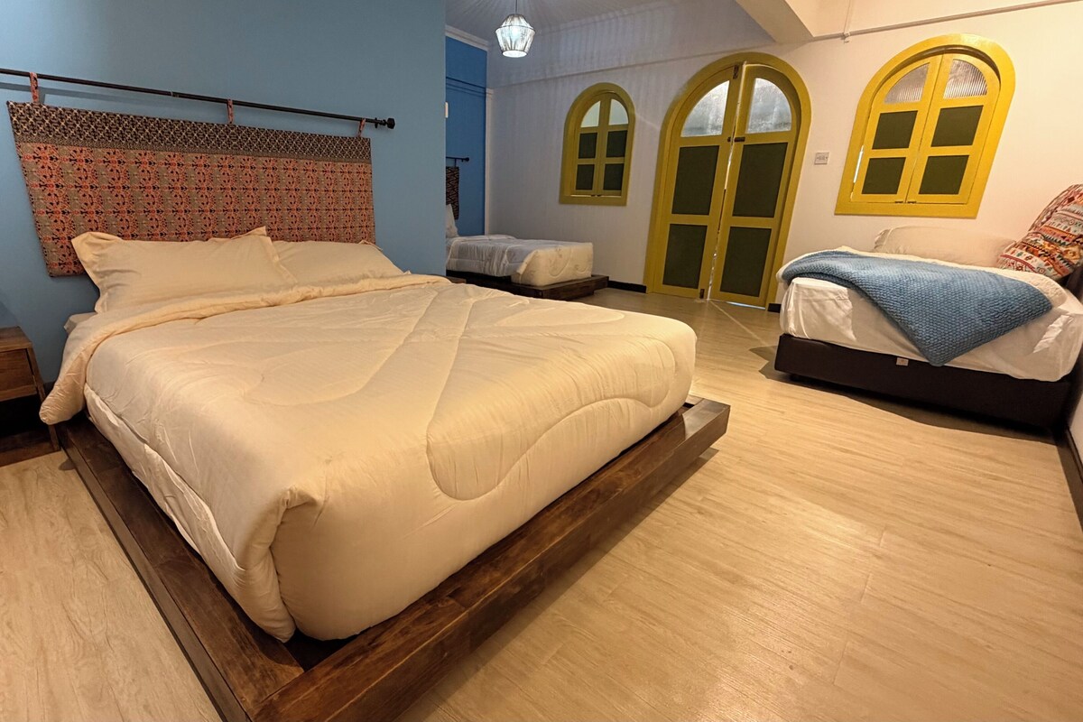 A bedroom with a large bed, a bedside table, a lamp, and two windows with yellow frames.