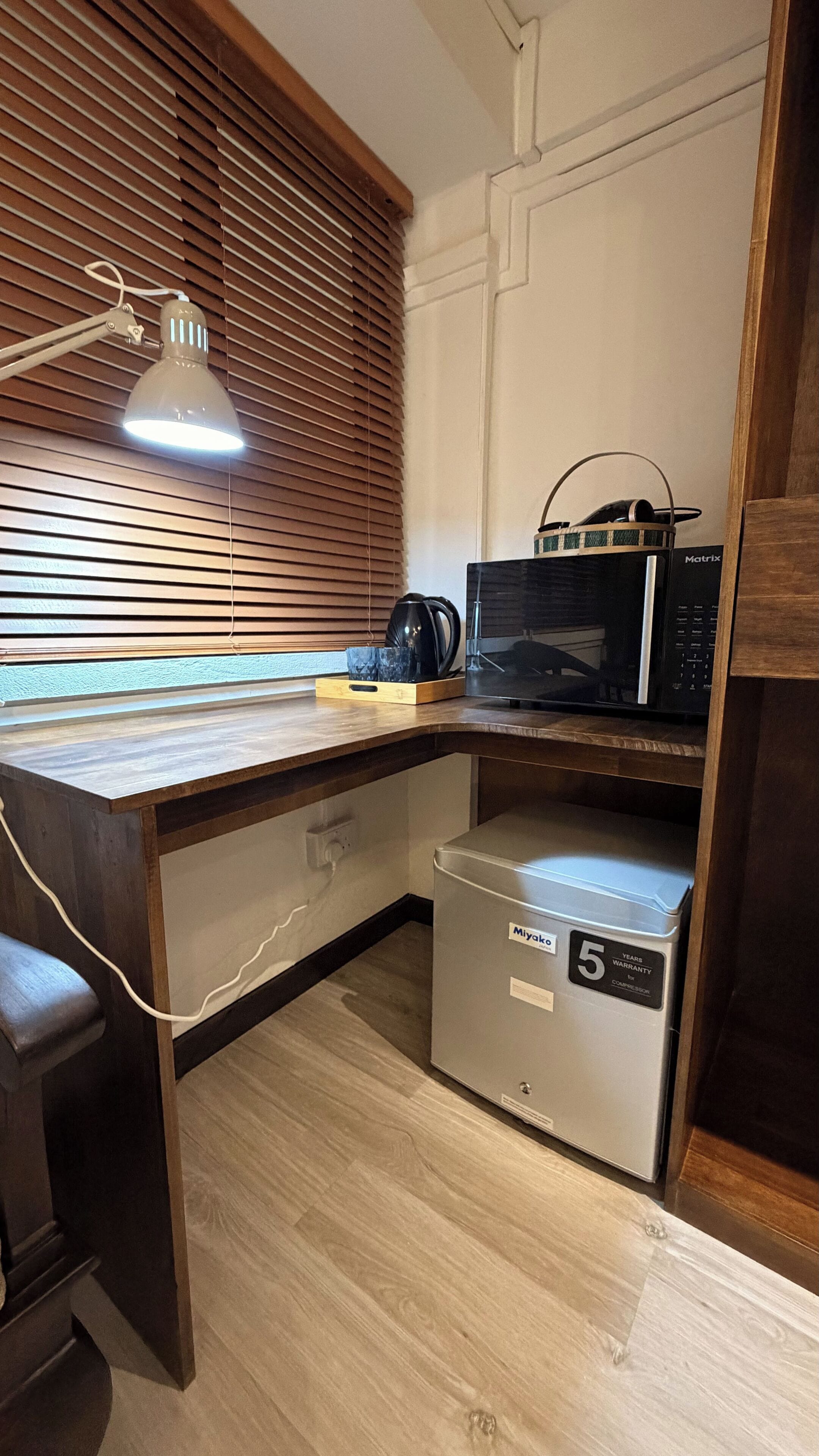A compact kitchenette with a microwave, a small refrigerator, and a wooden desk with a lamp.