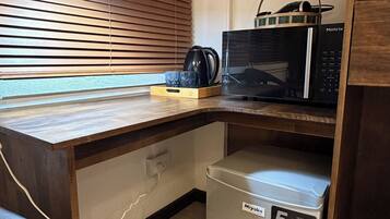 A compact kitchenette with a microwave, a small refrigerator, and a wooden desk with a lamp.