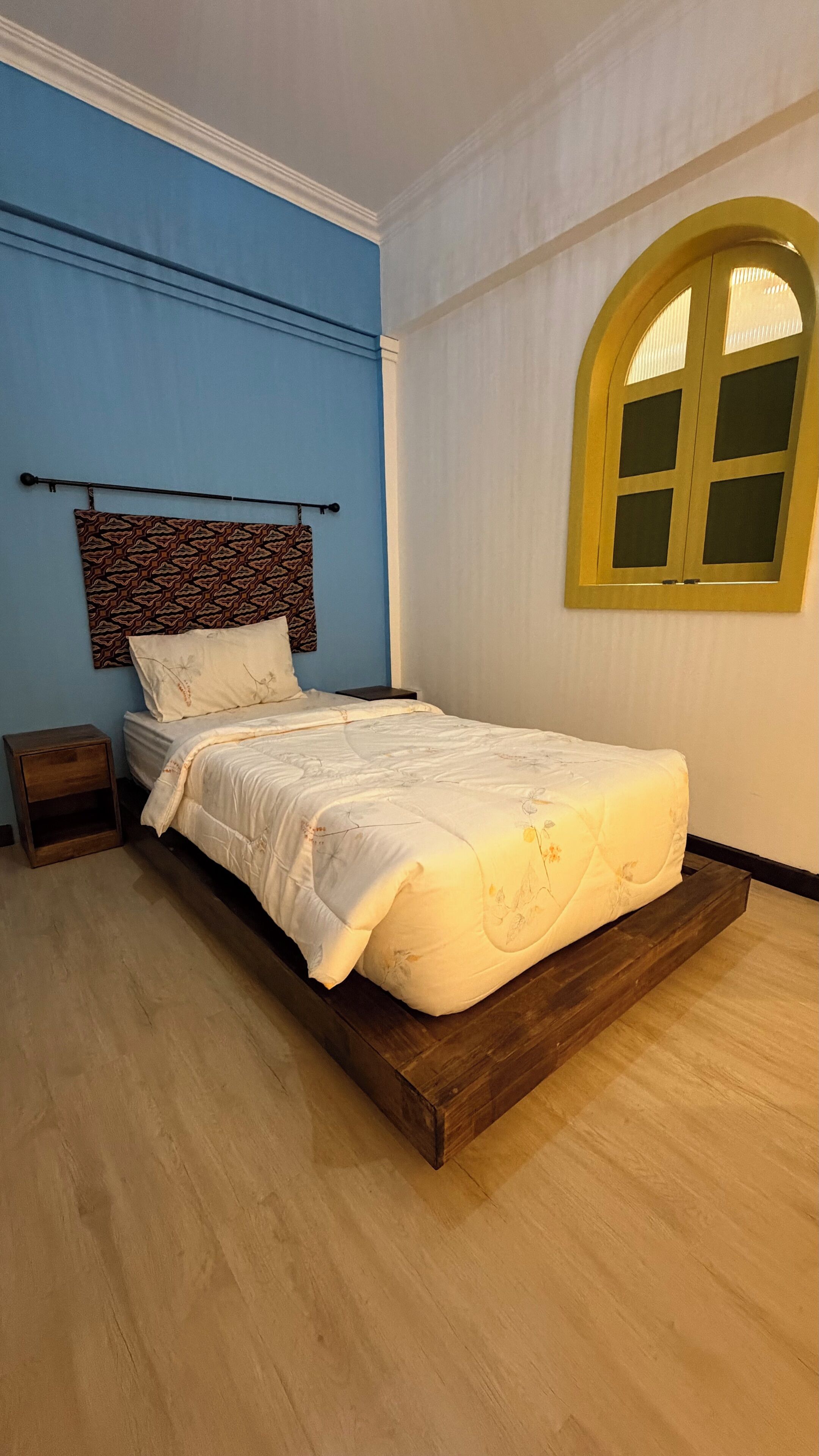 Une chambre à coucher avec un lit, une table de chevet, une tête de lit à motifs, une fenêtre avec un cadre jaune et un mur bleu.