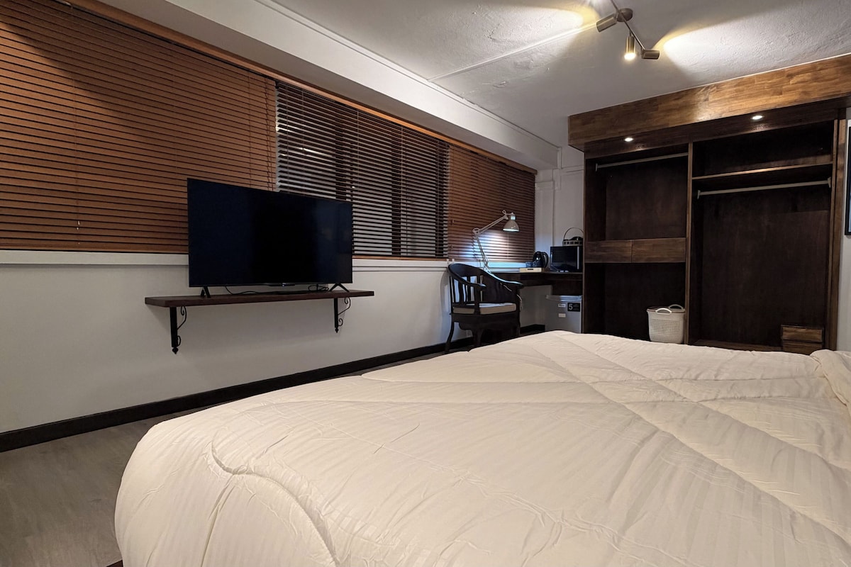 A bedroom with a bed, a television on a shelf, a desk with a chair, and a wardrobe.