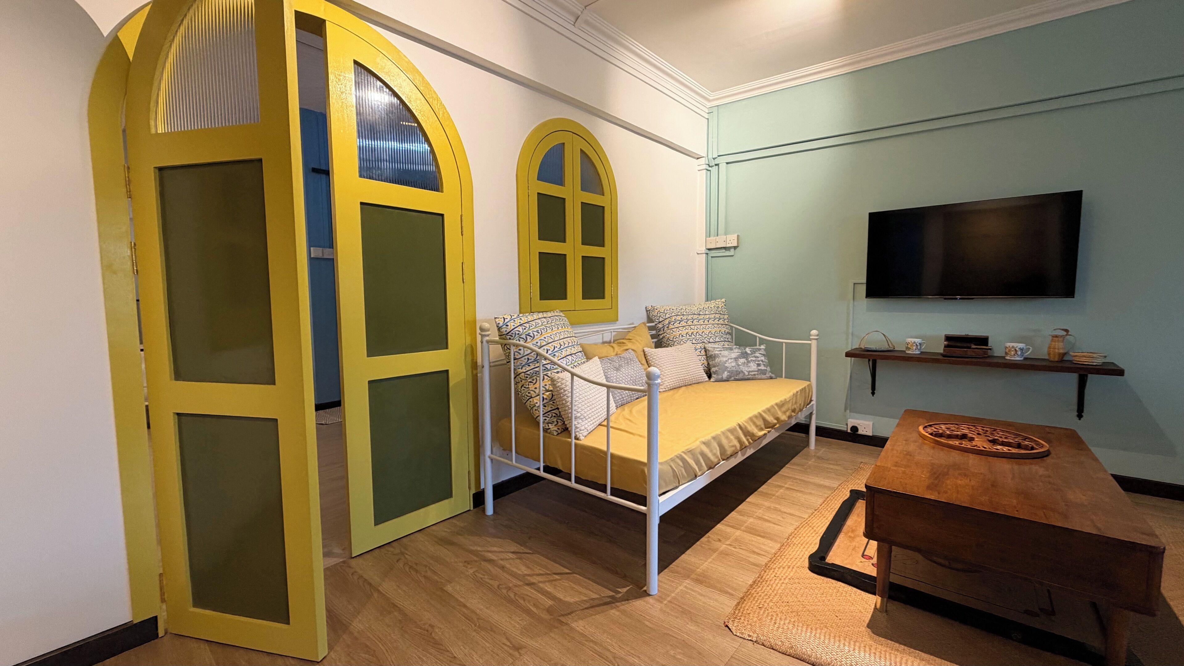 A room with a bed, a television, a wooden table, and a unique yellow arched doorway.