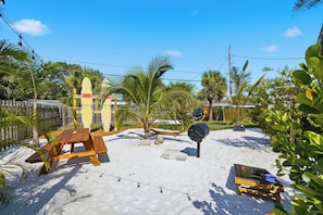 A sandy outdoor area with picnic tables, a barbecue grill, and surfboards labeled 'RESCUE' against a wooden fence. - Cocoa Cove: Steps to Beach, Walk to Downtown, Tiki Backyard (Cocoa Beach)