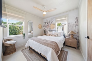 A bedroom with a bed, a nightstand, a chair, a ceiling fan, and a window with a view of greenery. - Cocoa Cove: Steps to Beach, Walk to Downtown, Tiki Backyard (Cocoa Beach)