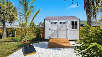 A small shed with a wooden deck in front, surrounded by palm trees and a grassy yard.