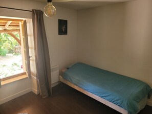 Un llit individual amb un mantell blau, una finestra amb cortines, un quadre penjat a la paret i llum penjant.