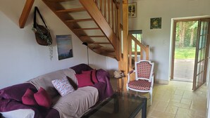 A living room with a sofa, a glass coffee table, a patterned armchair, a wooden staircase, and a door leading outside.