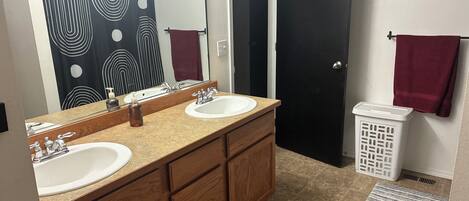 A bathroom with two sinks, a mirror, a towel rack, a laundry basket, and a patterned shower curtain.