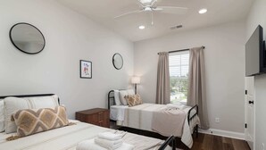 Ein Schlafzimmer mit zwei Betten, einem Deckenventilator, einem Fenster mit Vorhängen, einer Nachttischlampe und einem Flachbildfernseher.