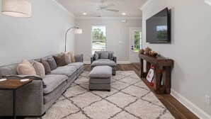 Ein Wohnzimmer mit grauem Sofa, Hocker und Couchtisch. Ein Fernseher ist an der Wand montiert und ein Deckenventilator vorhanden.